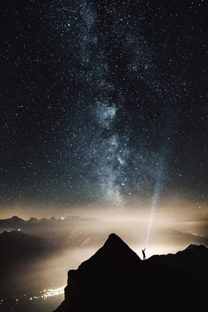 A man on a mountain gazing up at the stars, shining a flashlight beam into space